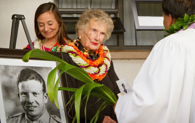 Elderly woman wearing lei with photo of young soldier outdoors facing minister with ti leaf