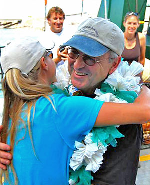 Smiling man with ball cap and large paper lei hugging a woman