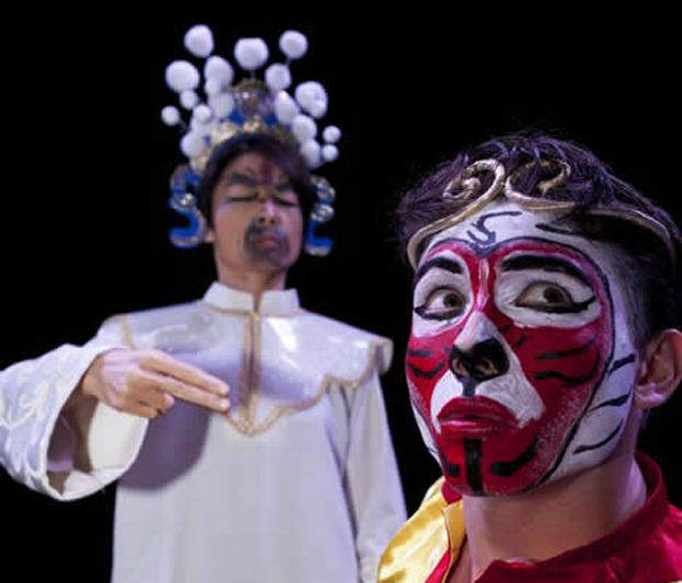 one actor in a white robe with elaborate crown and another, looking away in a bright red monkey mask