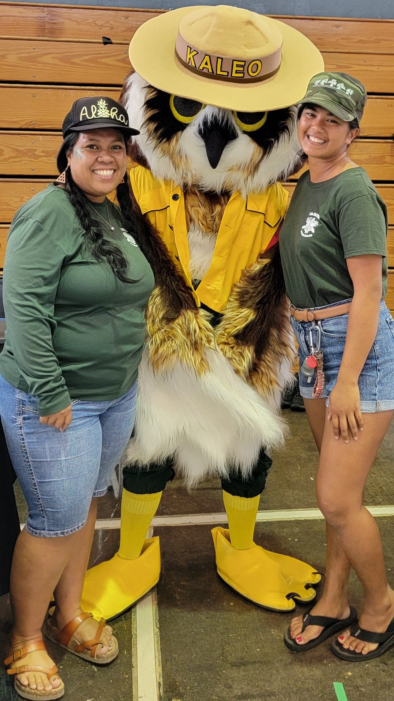 Two young women stand on either side of a person in a costume of a Pueo wearing a hat.