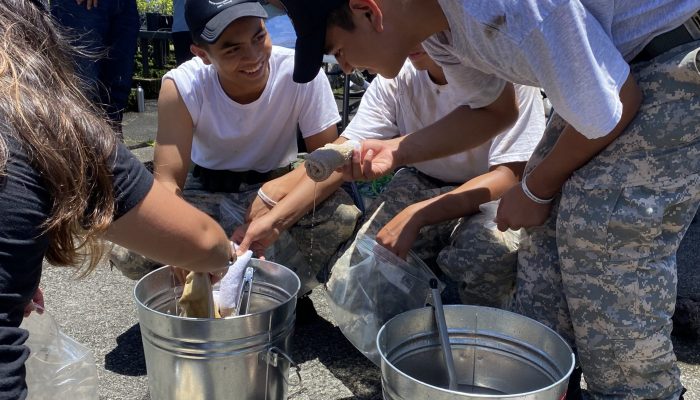 Three boys gather around two large buckets, placing fabric into them to dye it.