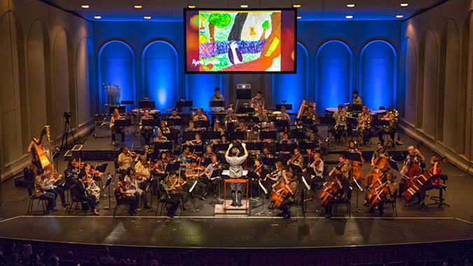 Wide shot of the symphony performing with animation on a screen above the stage