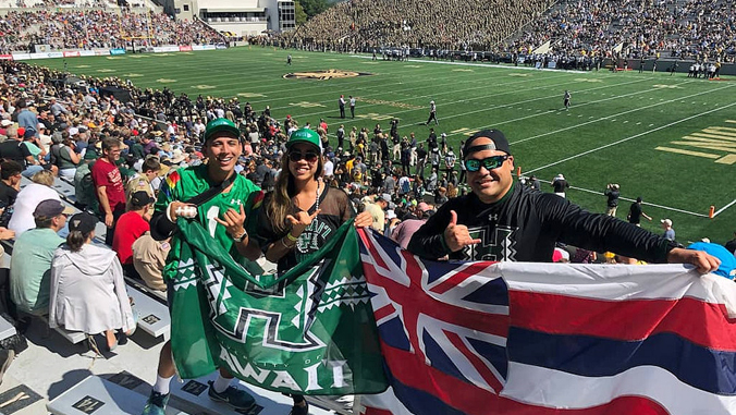 two people holding UH athletics flag and one person holding Hawaiian flag at West Point stadium