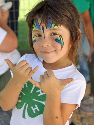 girl with face painted wearing 4-H shirt and waving shaka
