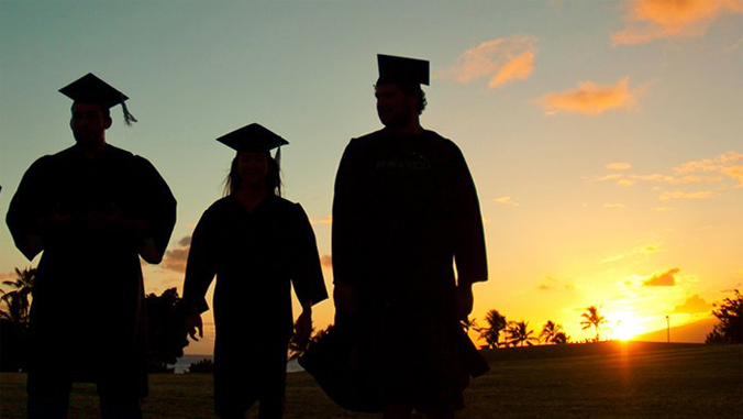 3 students in commencement gown with sunset behind them