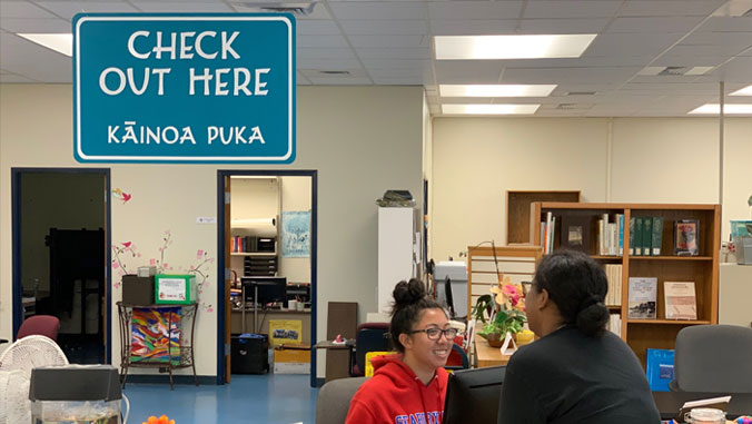 Two students talking in U H Maui College's library with a sign that says Check Out Here and the words in Hawaiian