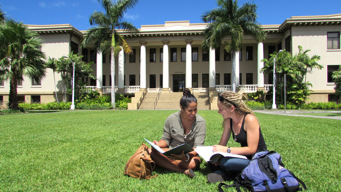 two people looking at a book and stiting on grass in front of a building