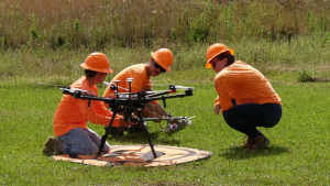 Three people with protective helmets and high-visibility shirts work on a drone in a field
