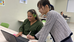 Student seated at a desk with a laptop and an instructor standing nearby