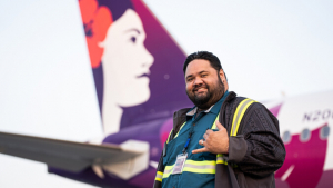 Man flashing shaka in front of a Hawaiian Airlines plane