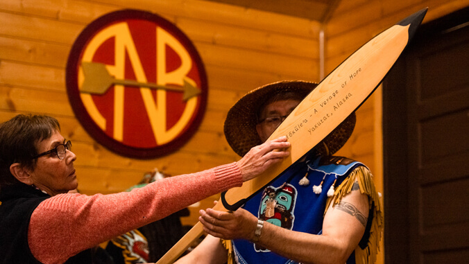 Two people holding a wooden paddle that contains the text: Hokulea a Voyage of Hope Yakutat, Alaska